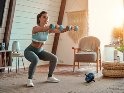 athletic girl in leggings and top crouches with dumbbells at home.