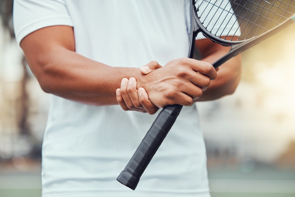Closeup of unknown indian tennis player suffering from wrist injury in court game. Ethnic fit professional in pain while holding and rubbing hand after match. Sporty man holding racket in sports club